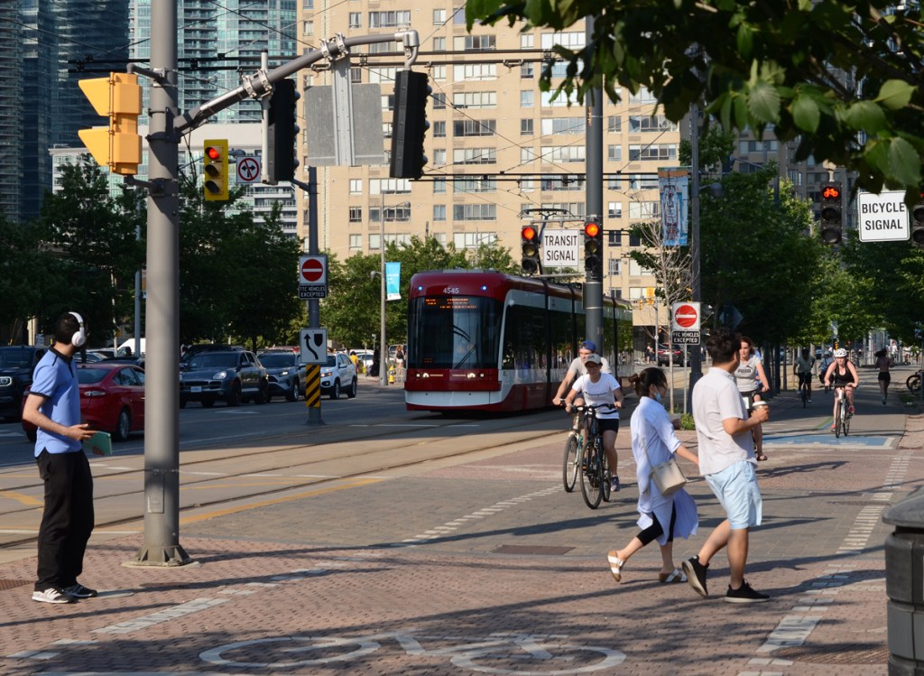 TTC street car on Queens Quay, cyclists on the bike path, pedestrians trying to cross