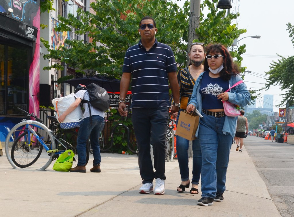 3 pedestrians walking on a sidewalk, walking past a man struggling with a large package on his bike
