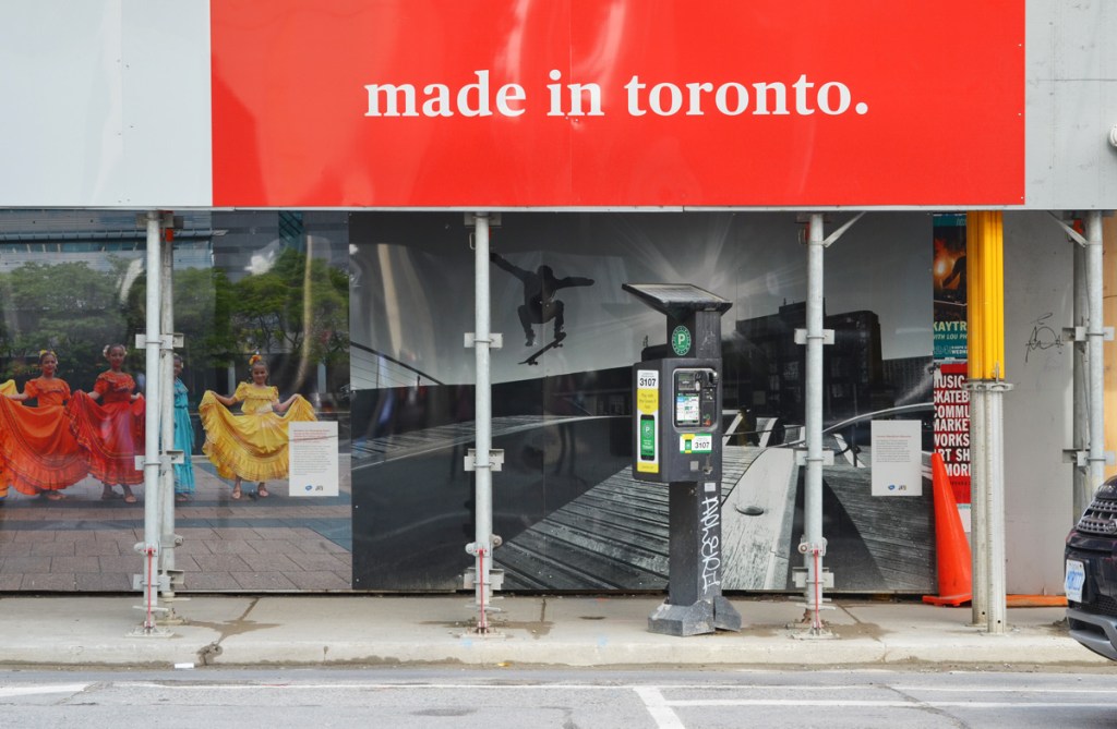 hoardings at a construction site, large red and white sign on top that says made in Toronto, a parking meter machine on the sidewalk, two large photographs on the hoardings, part of an exhibit 