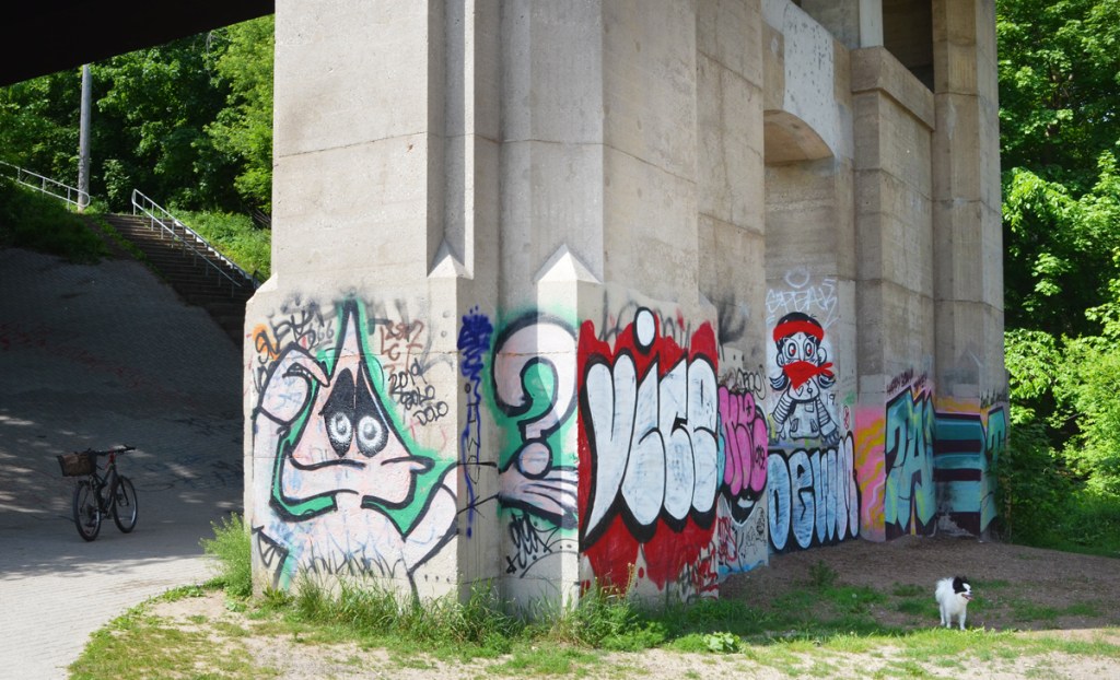 a small black and white dog stands beside graffiti on the bottom part of a bridge support. triangle headed man holding a big question mark, the word vice, a man with red head band and red bandana over lower part of face, 