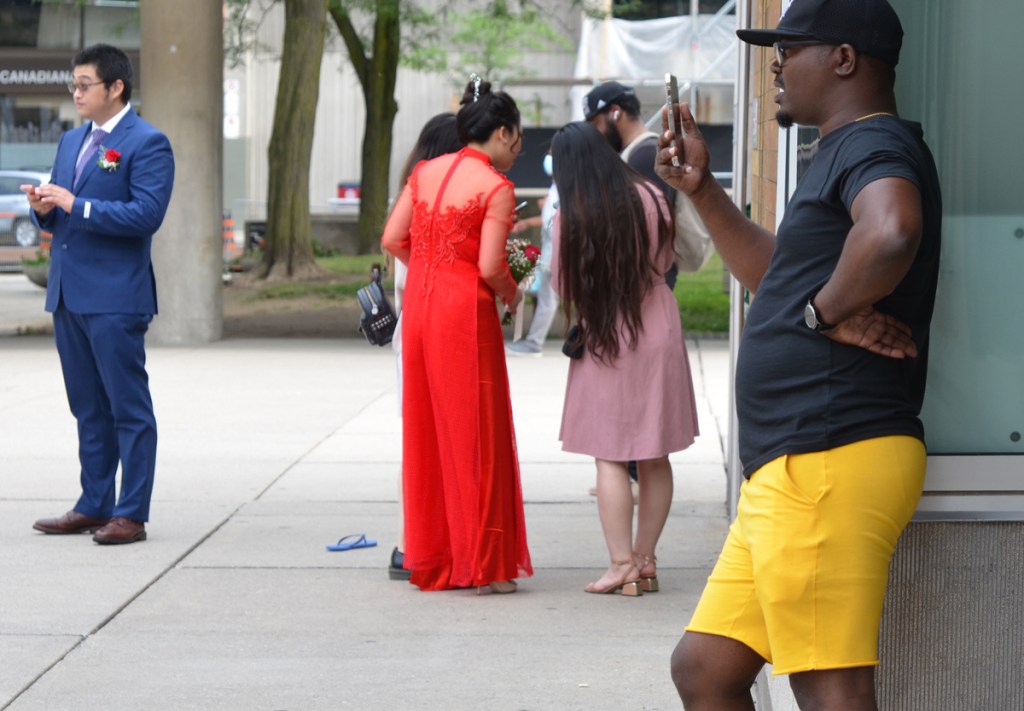 a black man in black baseball cap and T shirt and bright yellow shorts is taking photos with his camera. in background is a wedding shoot of Chinese bride in a long red dress
