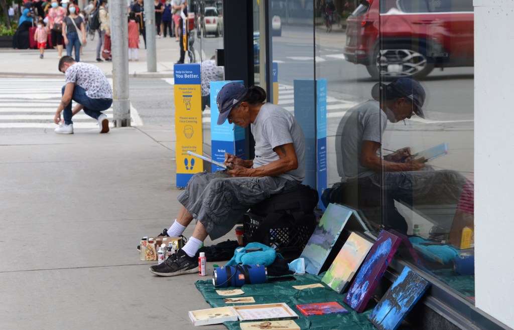 a man is drawing while sitting on the sidewalk, he has paintings and other artwork for sale, his reflection is in the window behind him. another man stops at the intersection to tie his shoe laces while waiting for the red light to turn green. Dundas Street