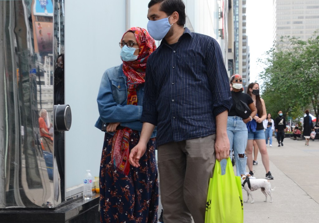 people waiting in line. first woman has mask, glasses and a red head scarf. she's with a man carrying a yellow plastic grocery bag, other women in the line behind them, at Yonge and Dundas