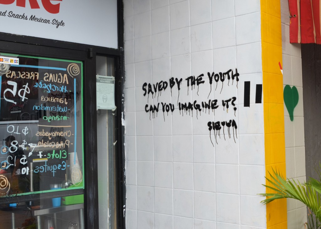 black letters spray painted on white tile wall, says saved by youth can you imagine it? wall is beside a window of a coffee shop with words and numbers listing the prices of drinks, aguas frescas