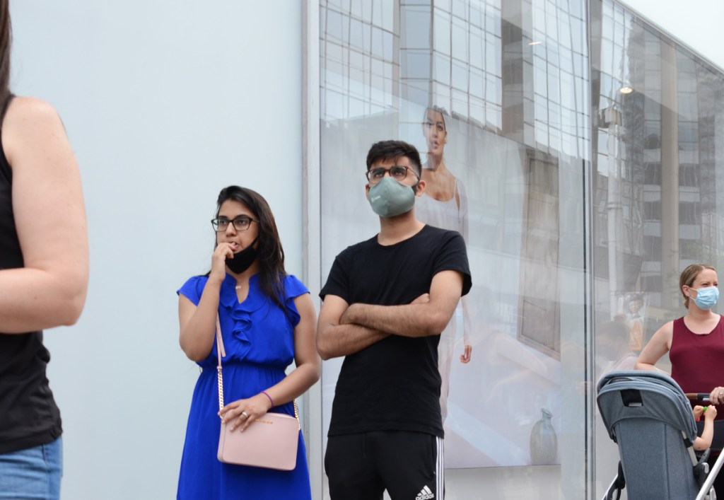 4 people in line to get into store at Yonge and Dundas, all keeping the 6 fet or 2 metres apart for covid restrictions. woman in red tank top is pushing a baby stroller, a couple stands together, woman in blue dress and black mask and a man in black, with advertisement behind him showing image of a woman in a white dress