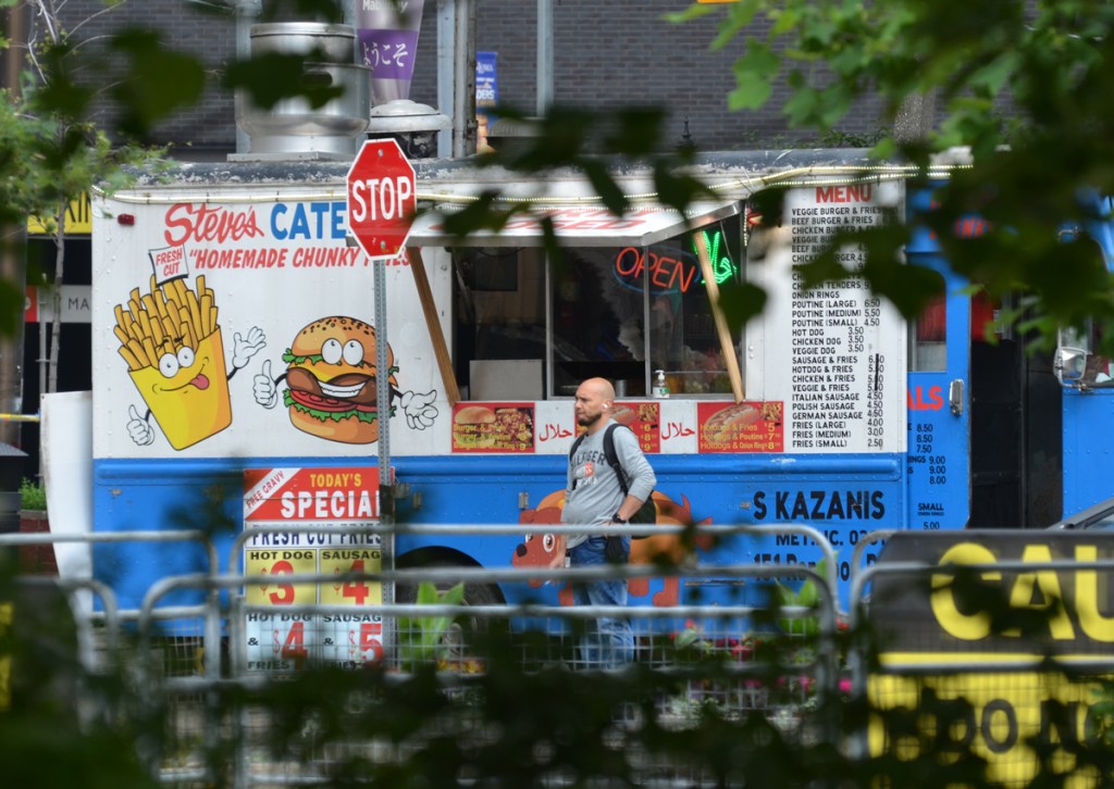 a man waits for his order from a food truck Steve's catering with images of cartoon fries and hamburger on the side of the truck, S. Kazanis truck, menu also on side of truck, today's special hot dog three dollars and sausage four dollars