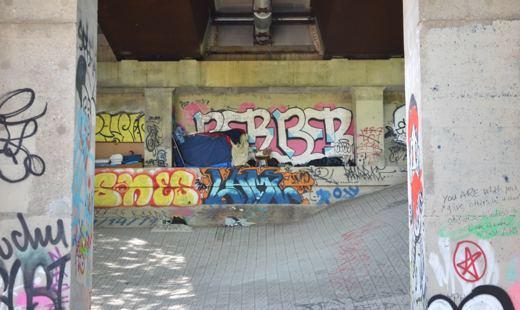 tent and belongings of a homeless man under a bridge surrounded by tags and other graffiti 
