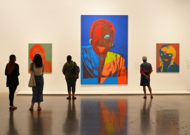 people standing in an art gallery looking a three large and colourful paintings by Andy Warhol of faces
