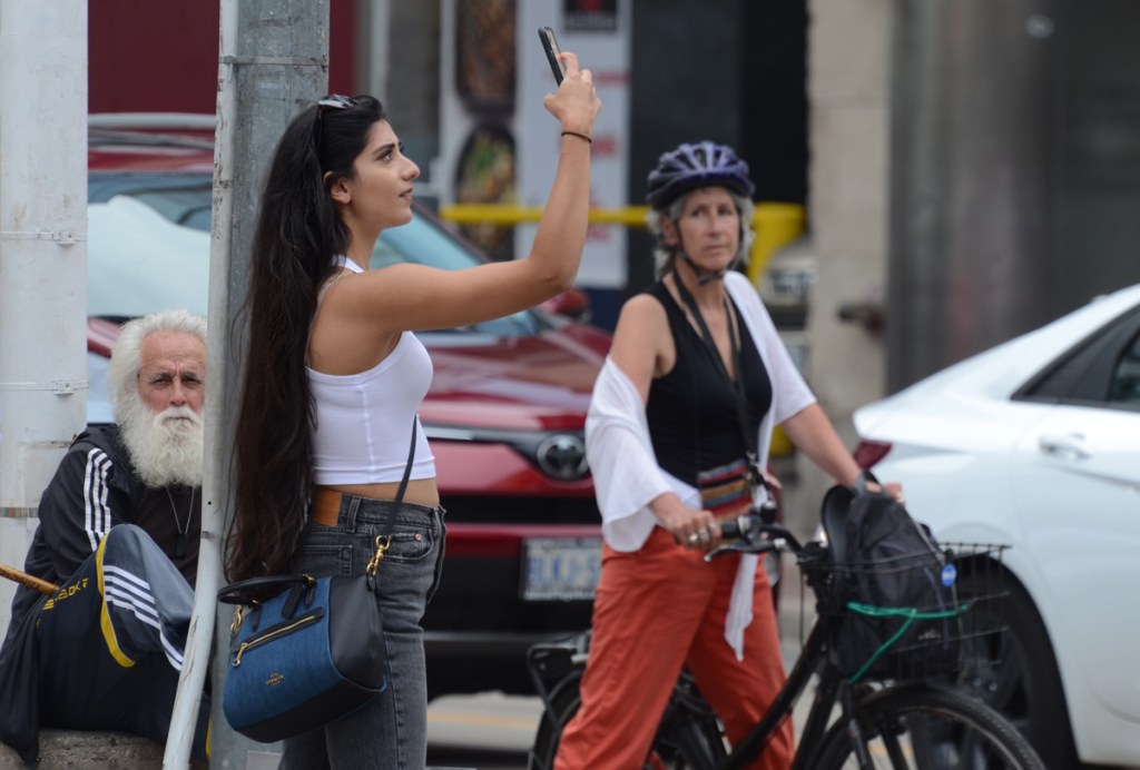 three people at Yonge and Dundas. One is a woman standing with her bike waiting for the red light, second is a woman in cropped white tank top taking a selfie, and third an older man with white hair, white beard and white mustache.