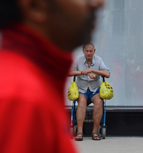 an older man in blue shorts and balding hair sits on a walker with a yellow grocery plastic bag on each handle. out of focus close up of man in red top in the immediate foreground