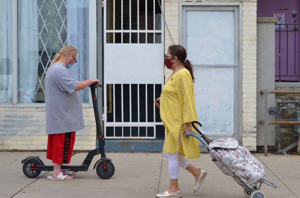 two people passing each other on a sidewalk, a man in red raptors shorts on a scooter and a woman with a long yellow top pulling a shopping buggy