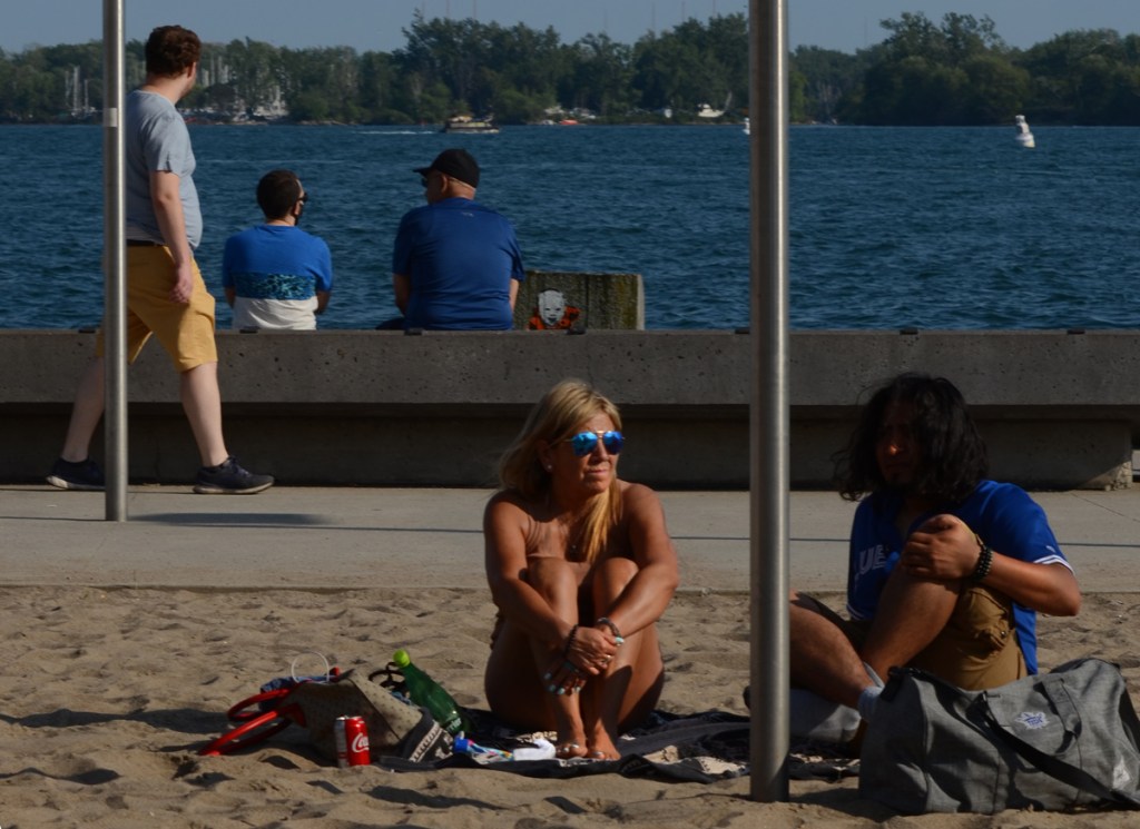 A couple sit on the sand on waterfront, under yellow umbrella, a man walks past behind them, also two men sitting on the edge of the lake,.
