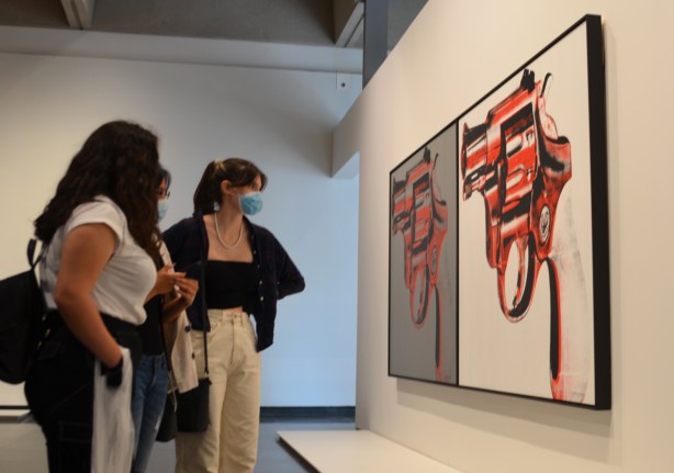 three young women look at two paintings of guns by Andy Warhol, large and on a gallery wall 