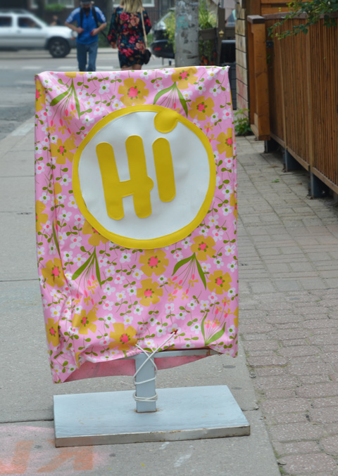 sign on sidewalk made with pink fabric covered with yellow and white flowers, a big white circle in the middle with hi written in large yellow letters
