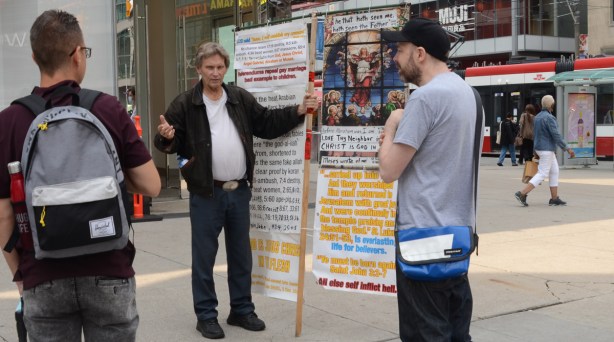 a man stands beside his Christian religious display at Yonge Dundas square while he talks to two men about Jesus 
