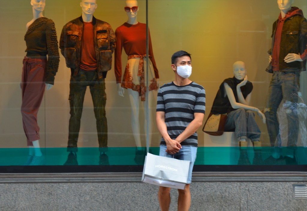 a young man with a mask and a Nordstroms shopping bag stands in front of the H B C windows with 5 mannequins modelling clothing