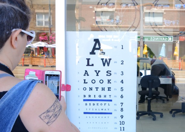woman taking photo with a phone camera of an eye chart in the window of an optometrist that reads always look on the bright side of life