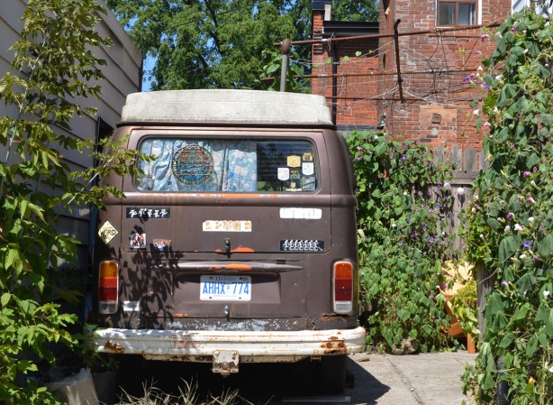 a brown van is parked in a backyard, an old green peace save the whales sticker is on the back window, blue and white curtain inside over same window, other old stickers on the back of the van