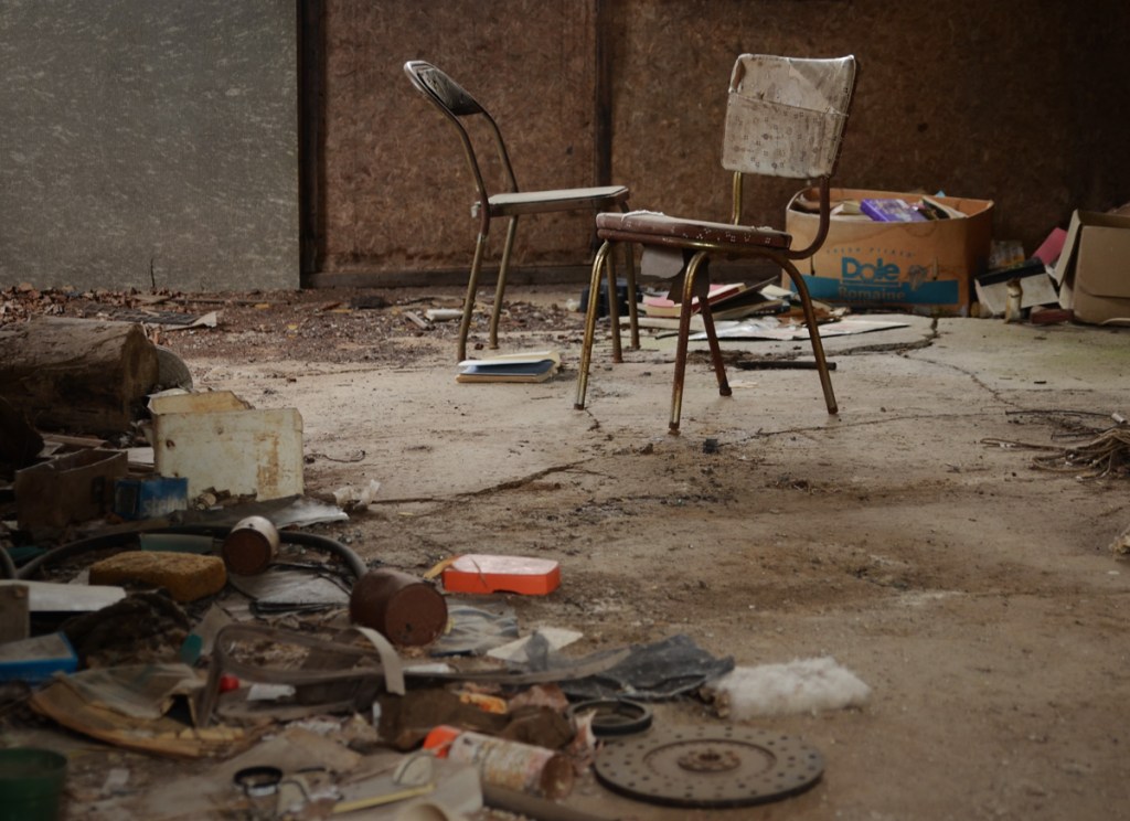interior of old shed with two chairs, debris on the floor and boxes of books in the corner