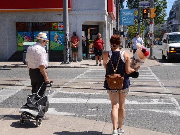 people waiting for a traffic light to turn green, and older man wearing a mask and pulling a shopping buggy, a woman carrying a boquet of flowers