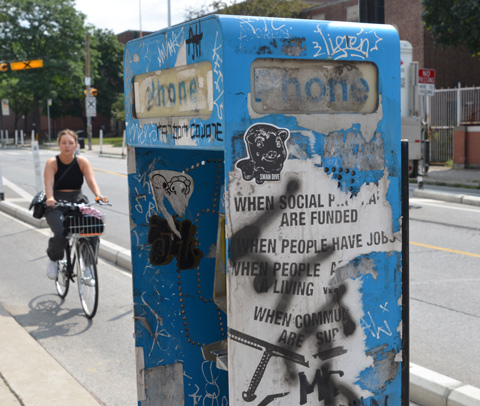 poster on a blue phone box that is torn and tagged, a woman on a bicycle is riding by