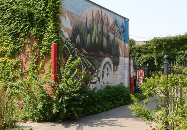 ivy on a wall of a building, a mural on the other wall, facing an alley. lake and mountains and trees in the mural