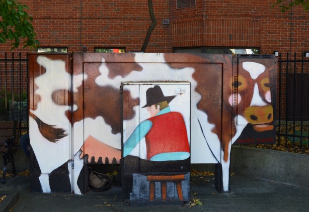 bell box on sidewalk painted to look like a cowboy on a small stool milking a brown and white cow 