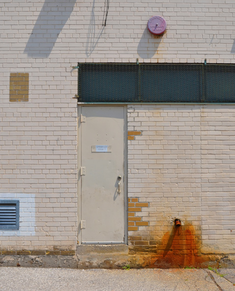 beige brick wall with beige metal door. sign on door says sprinkler room 7. rust marks on wall by overflow pipe 