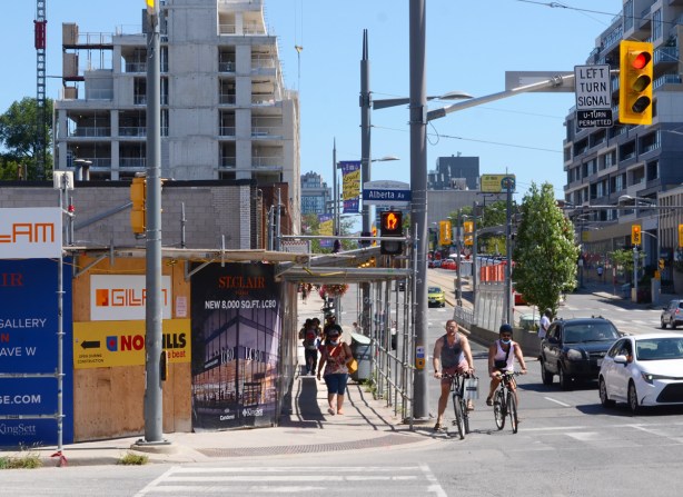 intersection of St. Clair West and Alberta, cyclists waiting for traffic light, hoarding around construction site on the northeast side, looking east