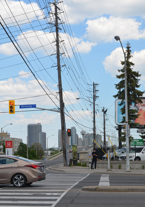 looking west on Eglinton at Kennedy, north side of the street, a man standing at the corner, hydro poles, high rises in the distance, traffic.