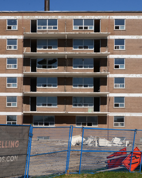 brick apartment building with balconies, empty, some broken windows, some white debris in a pile in front, blue construction fence 