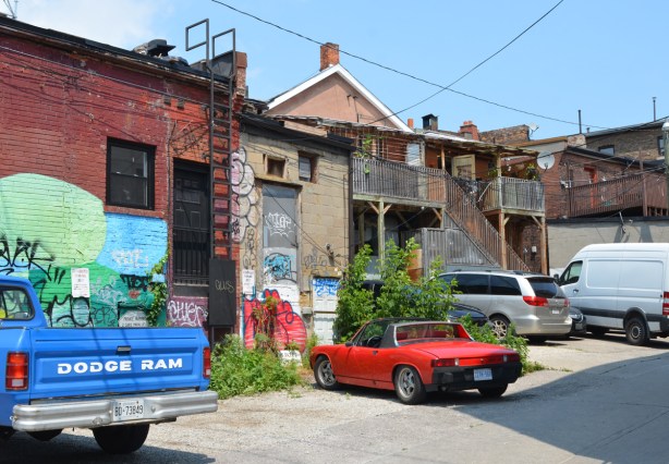 backs of stores in a small alley where a blue Dodge Ram pickup truck is parked. Also parked is an orange car, an old porsche convertible. Graffiti on some of the buildings 