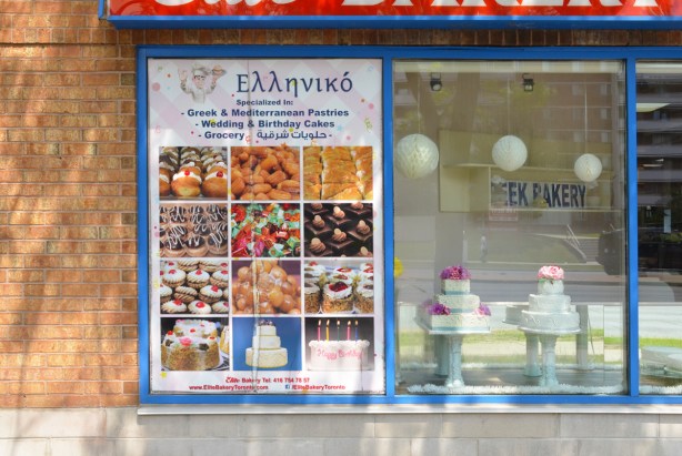 sign on the outside wall of a greek bakery, words in greek, anglish, and a south asian language (or arabic). wedding cakes in the window