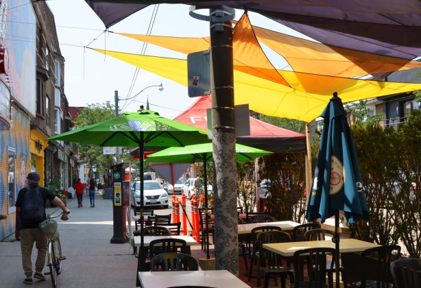 man walking on sidewalk, away from the camera. patio on street beside sidewalk with many umbrellas over the tables, green, red, and yellow