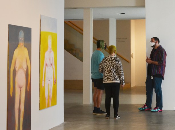 three people talking in a gallery with two large paintings of full frontal naked women on one of the walls