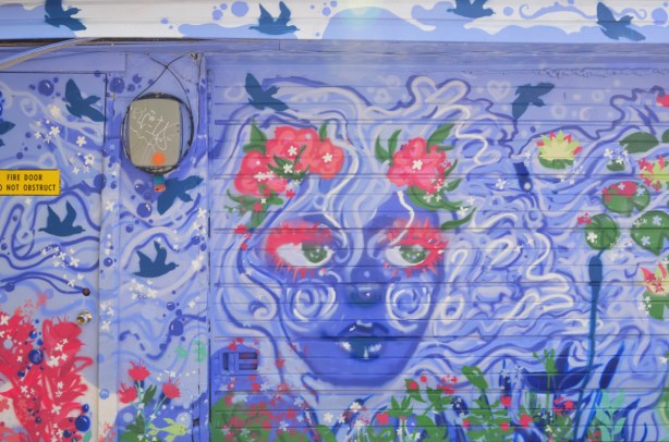 on a garage door in an alley, a mural of a blue faced women with swirly white and blue hair, flowers in her hair 