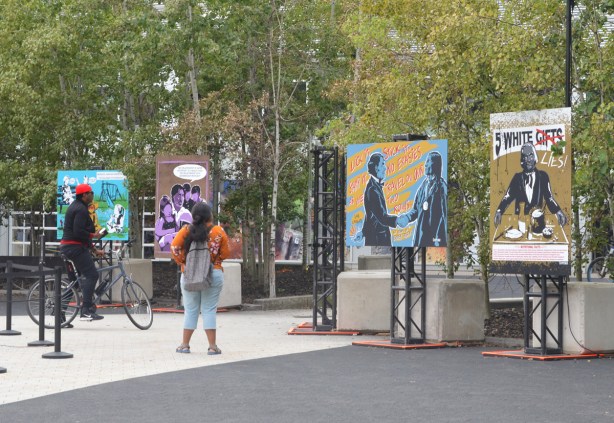 a man on a bike and a woman with a large backpack standing in front of posters by Jay Soule on display outside at Harbourfront, Indigenous Rights, history of abuse, protest, 