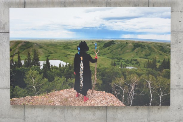 woman with back to camera on a rock ledge overlooking a green landscape large photograph by Meryl McMaster on display at Canada Square at Harbourfont