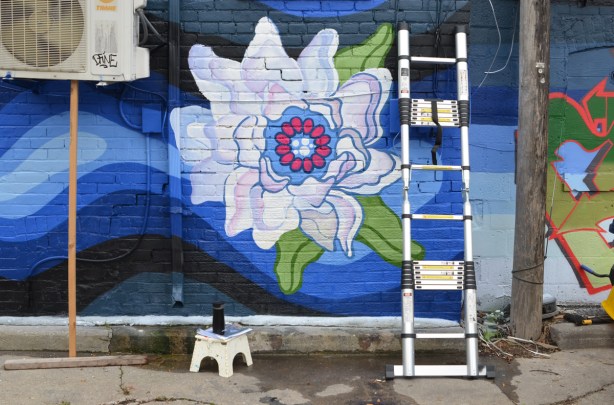 large white flower painted in a mural, on a blue watery background, ladder sitting in front of it, step stool with a can of spray paint