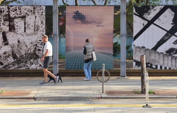 a woman stops to take a picture of a large photo on display on King Street West while a man and his black dog keep walking past the photos 