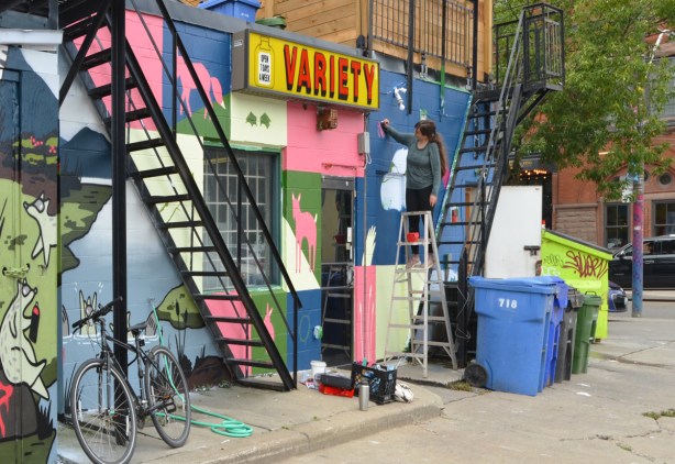 metal stair cases, exterior of a variety store, laneway, bike, garbage bins, artist on a ladder painting a mural