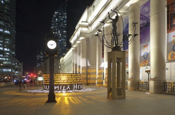 well lit Sir John A Macdonald Plaza in front of Union Station, at night, with lit clock, sculpture and an art installation by Masai Ujiri