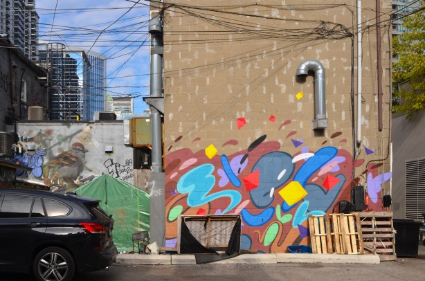 back of buildings in an alley, a mural of coloured blobs on the back one building, a small green tent, a picture of a sparrow on the other building, condos in the background