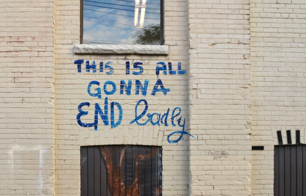 blue painted graffiti words on a pale grey brick building that say this is all gonna end badly