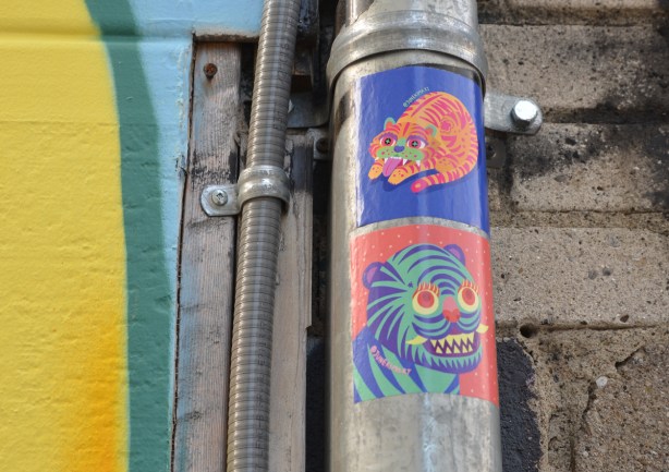 2 street art sticker slaps on a metal pole. both are images by June Kim. A cat and a tiger