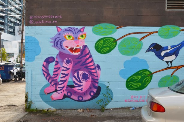 street art mural of a pink and purple striped tiger by June Kim, beside a tree with a blue bird in it (also painted)