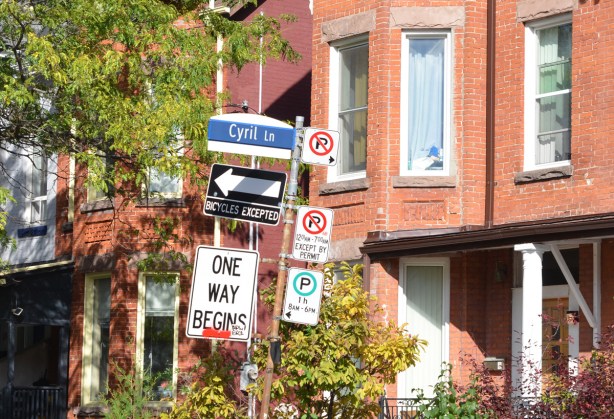 traffic signs on a pole in front of red brick rowhouses, Cyril Lane, plus no parking
