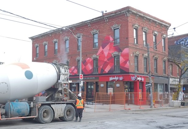 cement truck with a workman standing beside it, on Queen West, by a Wendys restaurant in a three storey brick building with a mural in red and black on the outside