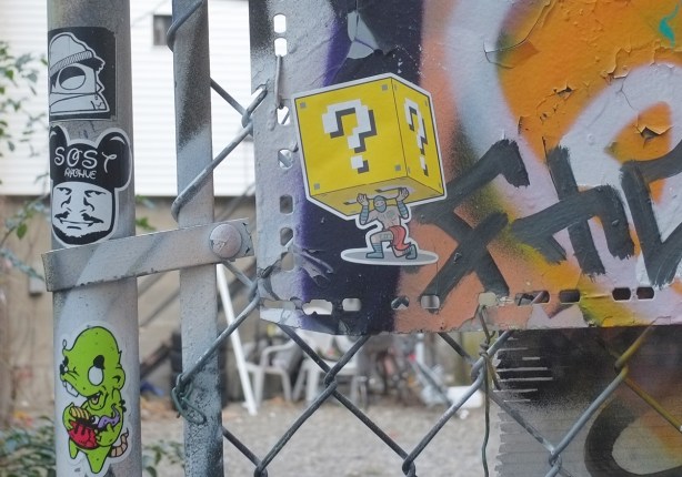 graffiti alley stickers including urban ninja squadron, t bonez on one knee while carrying a large yellow cube on his back. cube has white question mark on each face