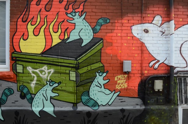 mural in an alley by Emily May Rose of raccoons and a big white rat. One raccoon is setting fire to a dumpster